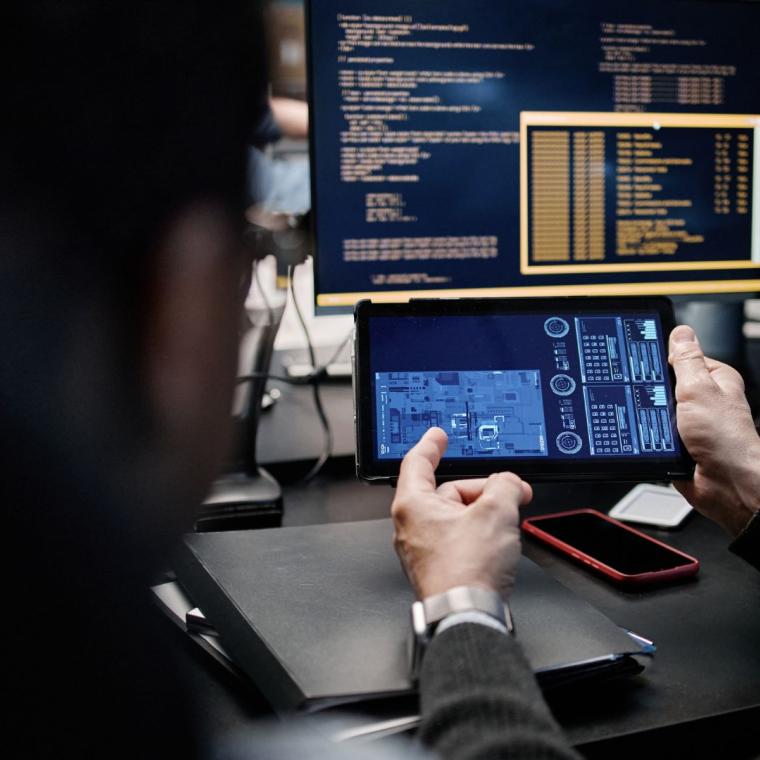 Middle aged Caucasian man holding digital tablet displaying technical schematics while sitting beside young man in front of computer monitors showing programming code