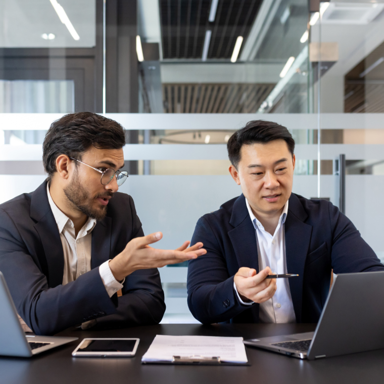 Two business colleagues discussing financial project while sitting at desk inside office, diverse people. Men serious and concentrated with laptops. 