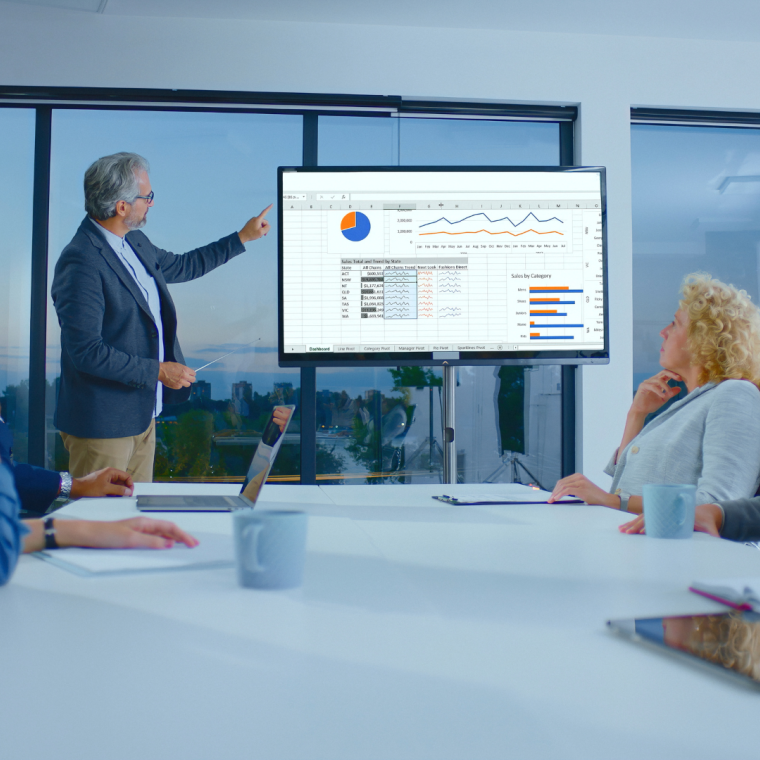 Senior businessman presenting financial data and charts on a large screen to a diverse team of colleagues participating in a corporate meeting in a modern office boardroom