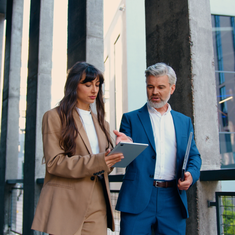 Confident business professionals in formal suits walking and talking outside a modern office building female executive using a digital tablet, male colleague holding a laptop