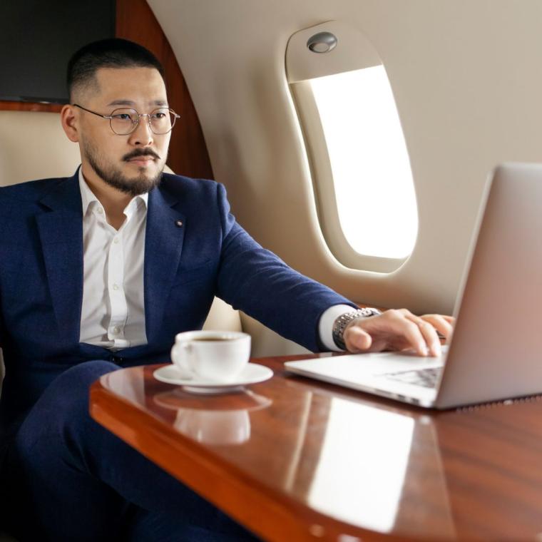 successful Asian businessman in suit and glasses flies in private luxury plane with laptop