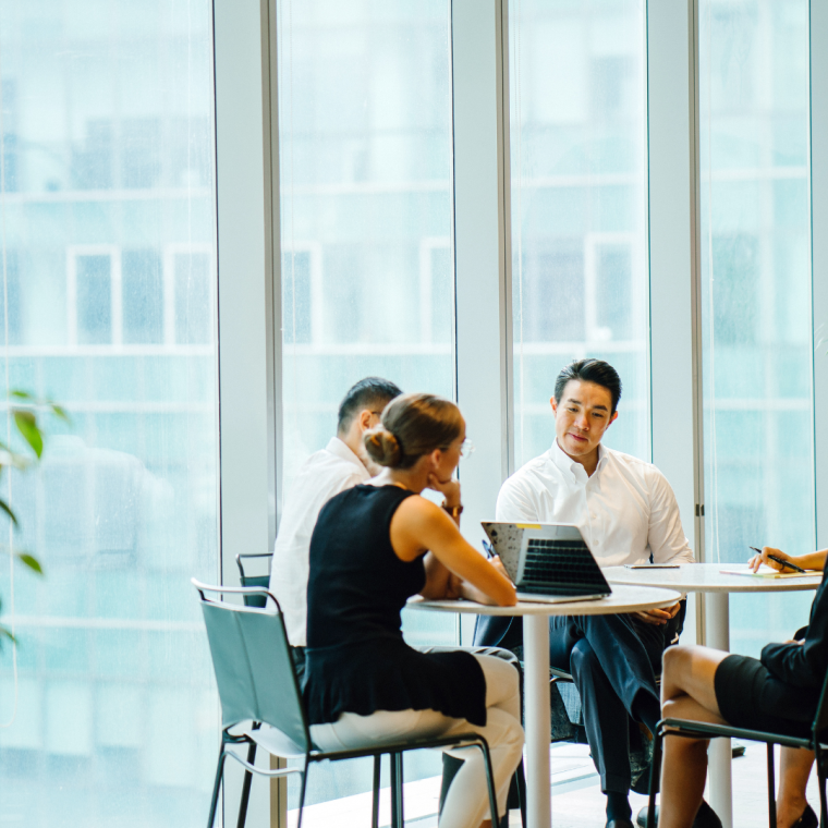 A diverse team sits around a table in a meeting room to have a business meeting to discuss plans. The group is international with Asian and white team members and they are all professionally dressed.