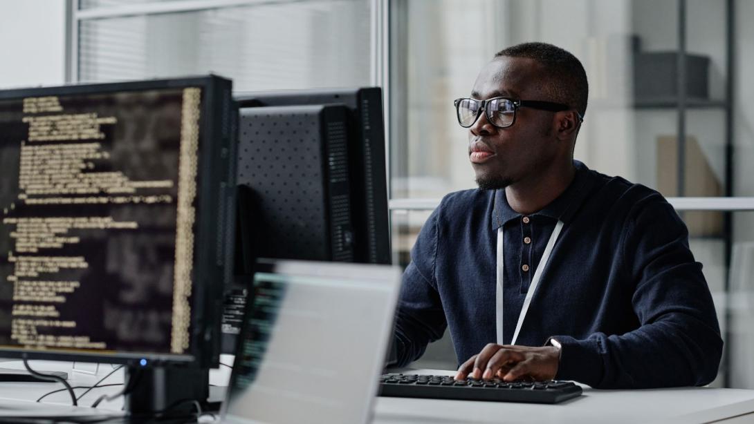 African American young developer in eyeglasses concentrating on his online work on computer sitting at workplace