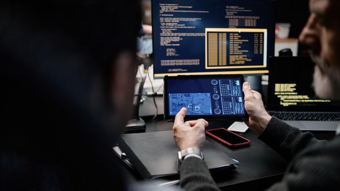 Middle aged Caucasian man holding digital tablet displaying technical schematics while sitting beside young man in front of computer monitors showing programming code