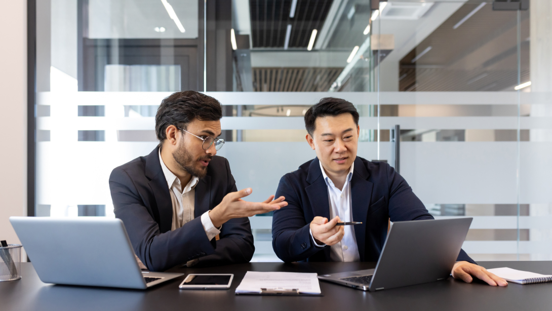 Two business colleagues discussing financial project while sitting at desk inside office, diverse people. Men serious and concentrated with laptops. 