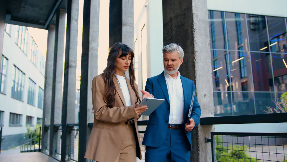 Confident business professionals in formal suits walking and talking outside a modern office building female executive using a digital tablet, male colleague holding a laptop