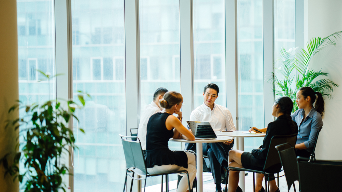 A diverse team sits around a table in a meeting room to have a business meeting to discuss plans. The group is international with Asian and white team members and they are all professionally dressed.