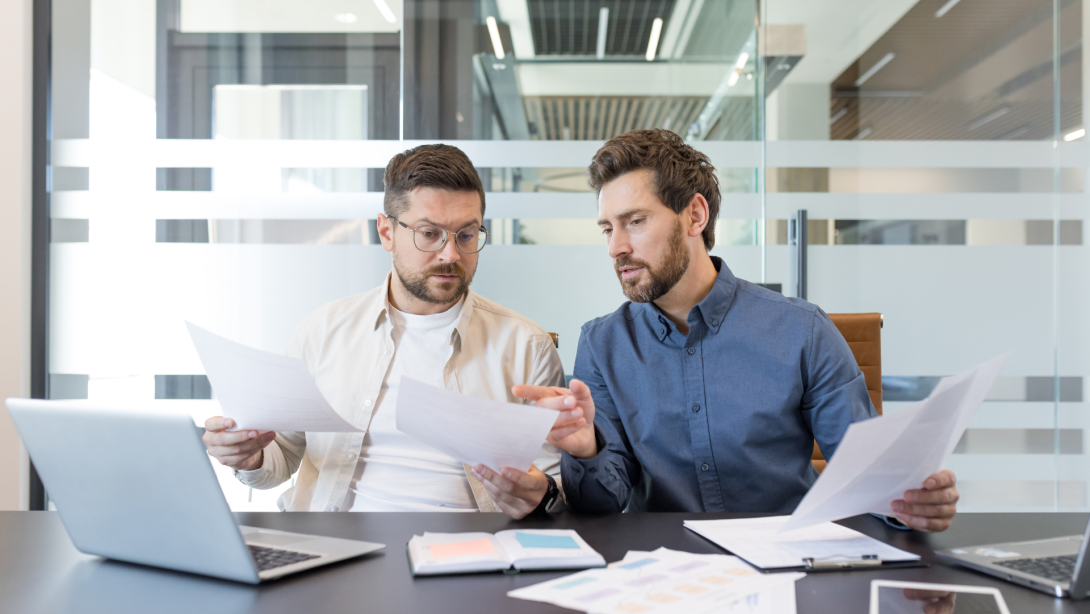 Two business colleagues working together, discussing documents and analyzing data during a corporate meeting in a contemporary office setting, demonstrating teamwork and partnership