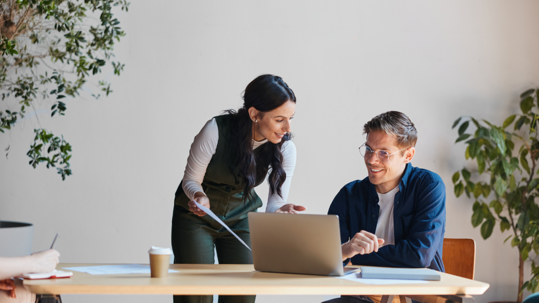 Business professionals collaborating at office desk, discussing project on laptop and documents, sharing ideas during meeting, working together in modern workspace