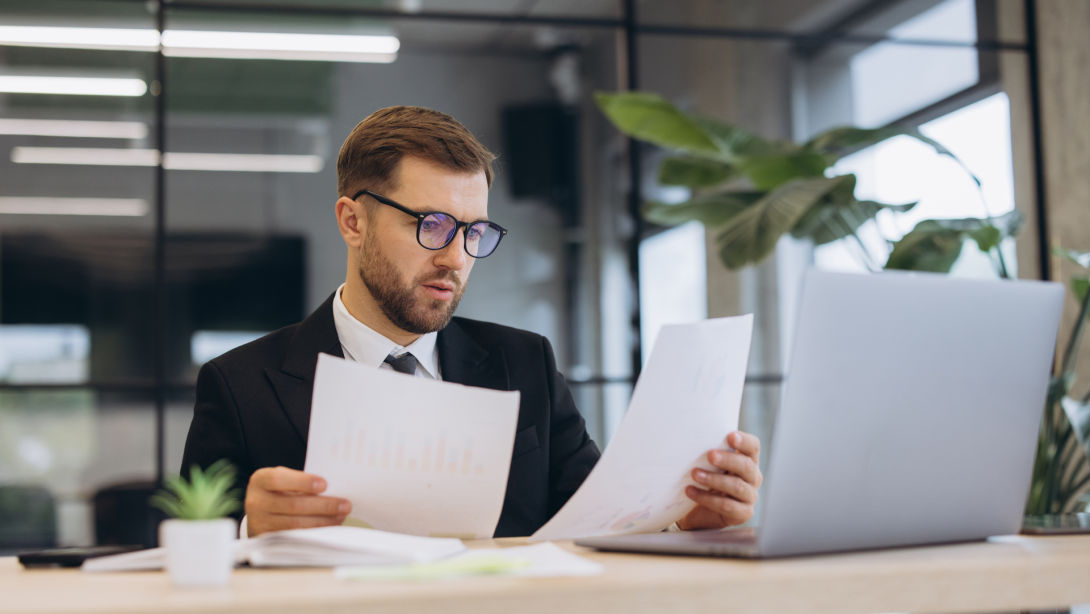 Young businessman reviewing financial reports in office, analyzing documents and working on laptop 